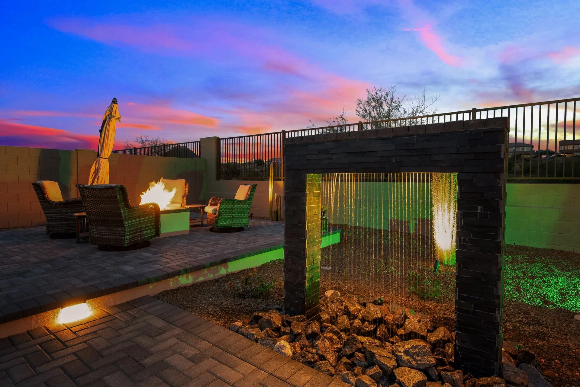 Backyard outdoor living design at sunset with a fire-pit lounge, water-wall feature, and accent lighting