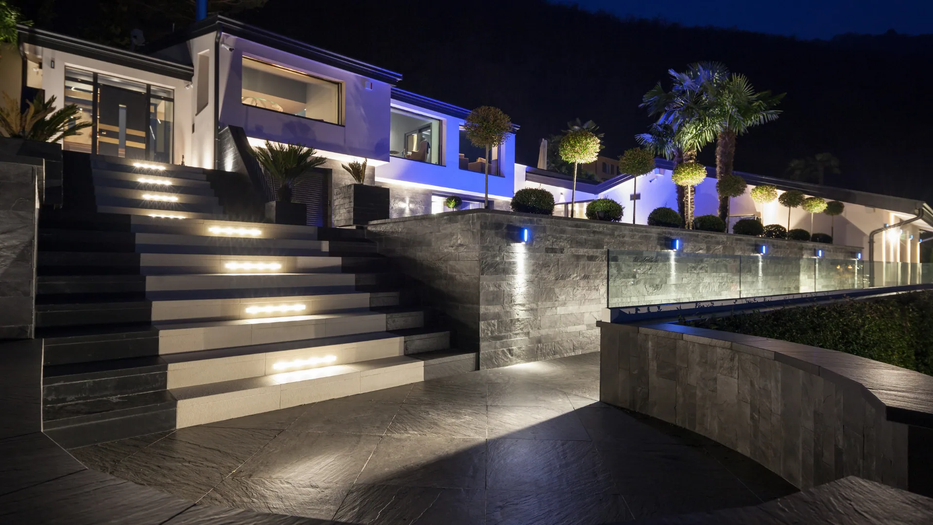 Modern home at night with illuminated steps, wall-wash, and uplit palms